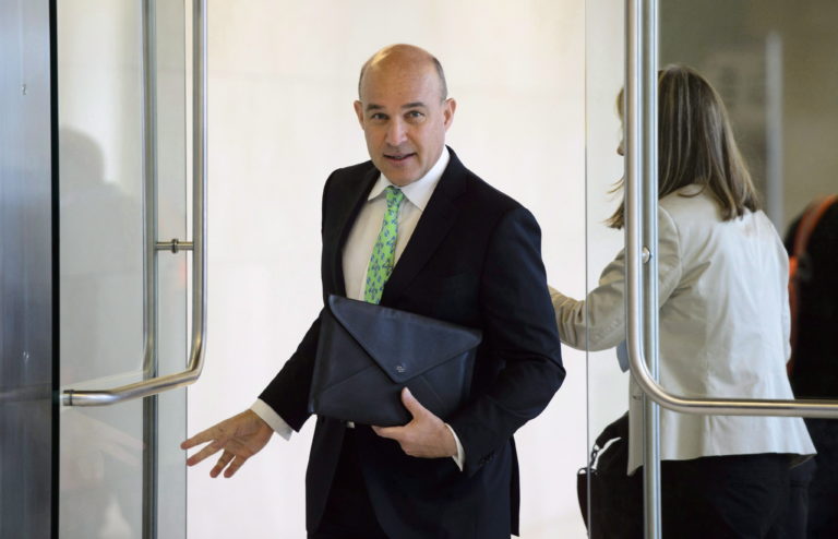 Jim Balsillie opens a glass door to enter a room; he's wearing a black suit with a bright, green tie and is holding a black folder for carrying papers.