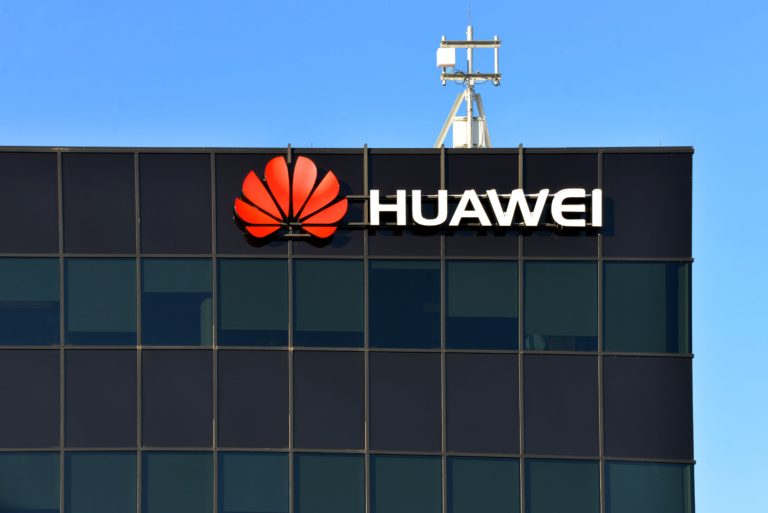 A shot of the Huawei logo on the top righthand corner of a black steel-and-glass office building
