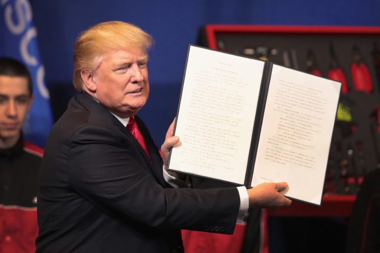 A head-and-shoulders shot of a smiling Donald Trump holding open a large black document cover bearing papers printed with a presidential executive order.