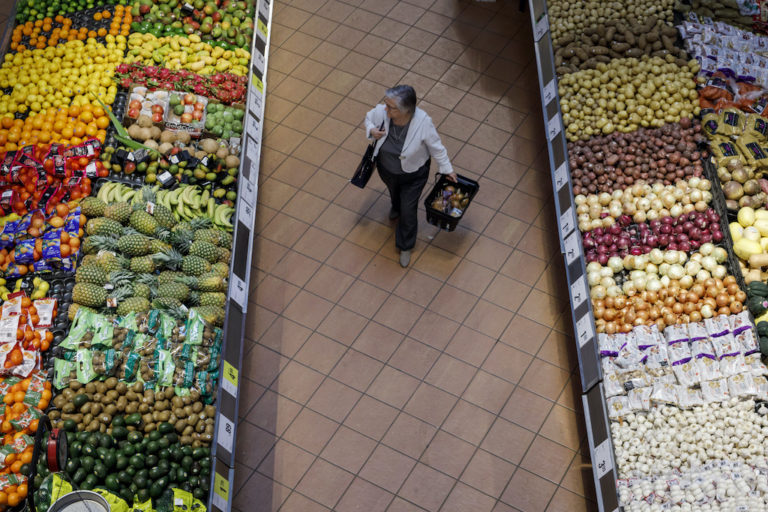An overhead shot of a woman walking alone down the middle of a wide produce aisle, with generously stocked counters full of fruit and vegetables on either side of her.