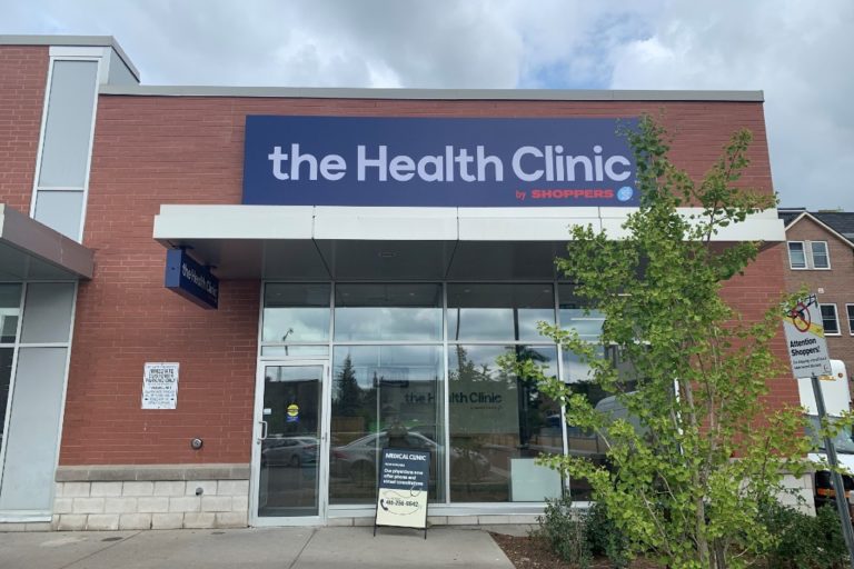 A modern red-brick building with large windows and a blue, tarpaulin-style sign that reads "the Health Clinic by Shoppers."