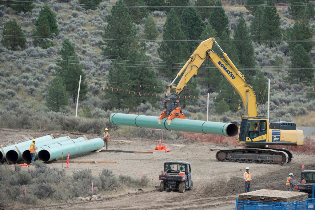 A wide shot of a pipeline construction area, with an excavator using its boom to lift a length of pipe.