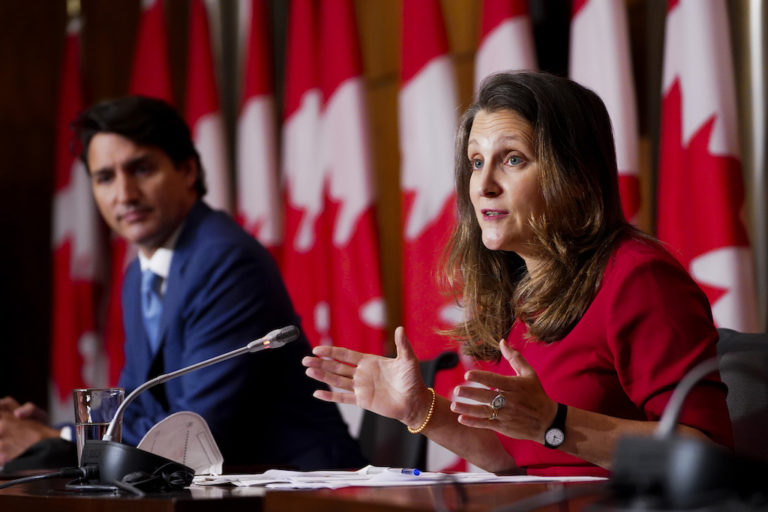 Chrystia Freeland and Justin Trudeau speaking at a press conference with Canadian flags in the background.