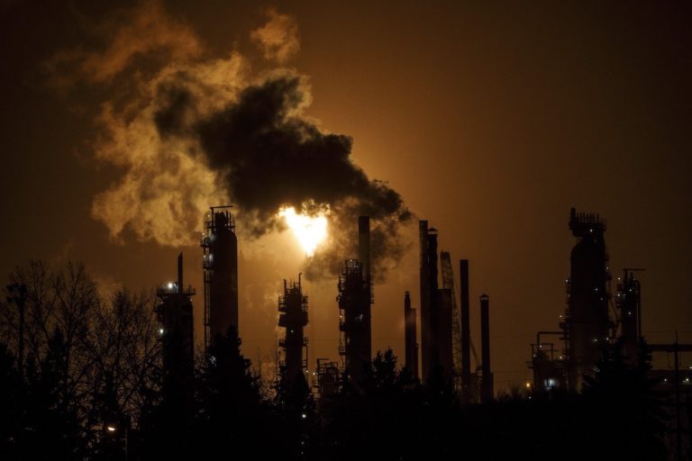 A silhouette shot of column and smoke stacks of a refinery. A bulb of flame is rising from one of the stacks, partially obscured by smoke from the others.