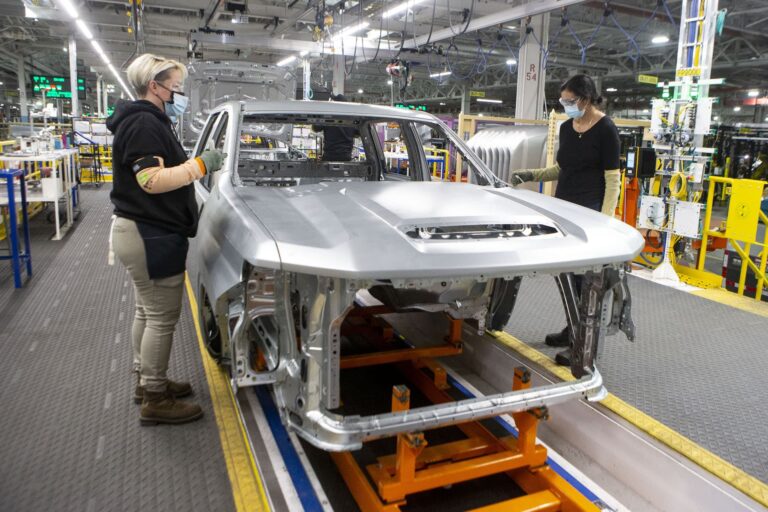 Two factory workers stand on either side of the silver-painted body shell of a pickup truck as it creeps down the assembly line.