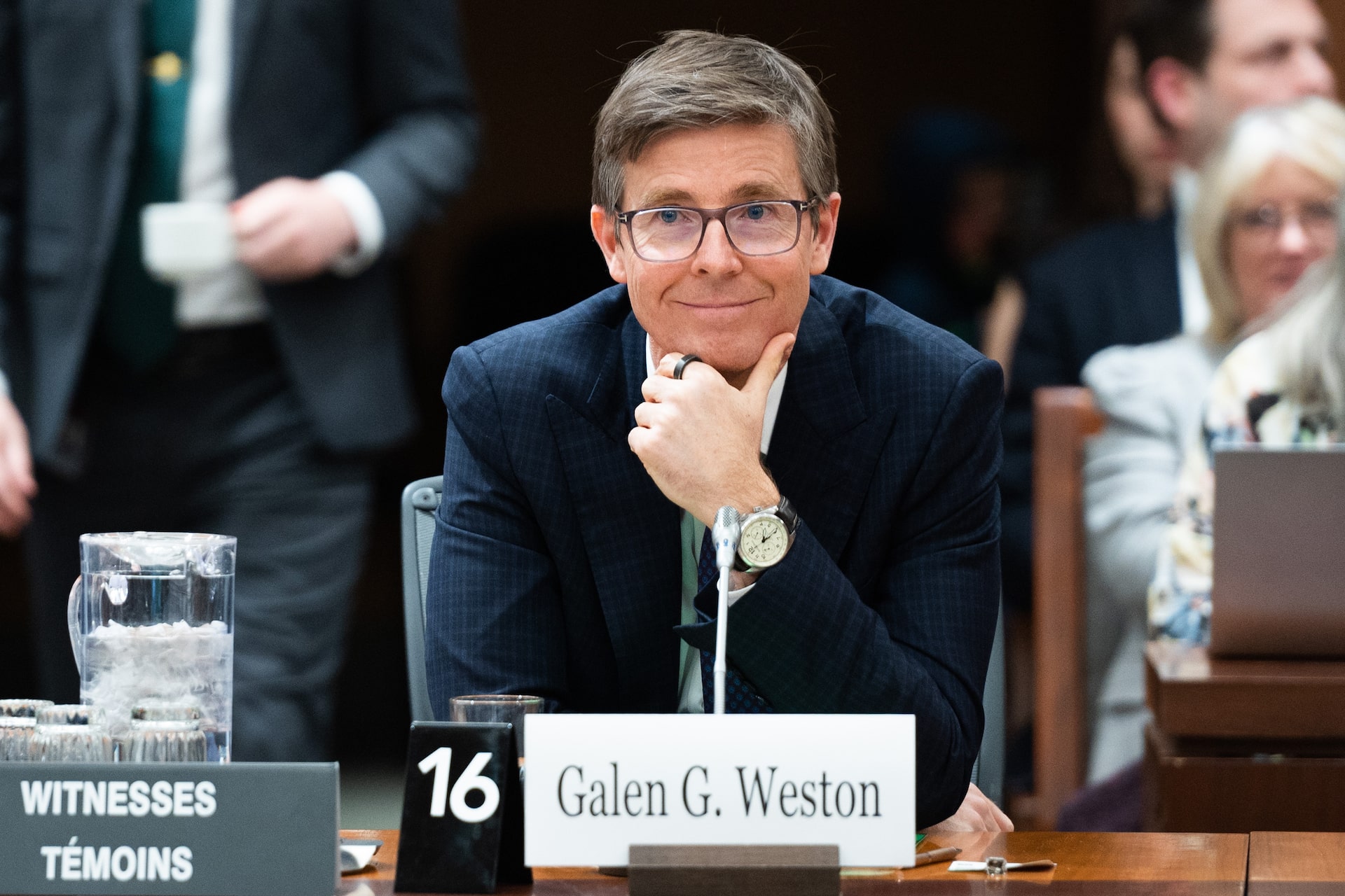Galen G. Weston, smiling and wearing glasses and a navy suit, sits at a Parliament committee hearing.