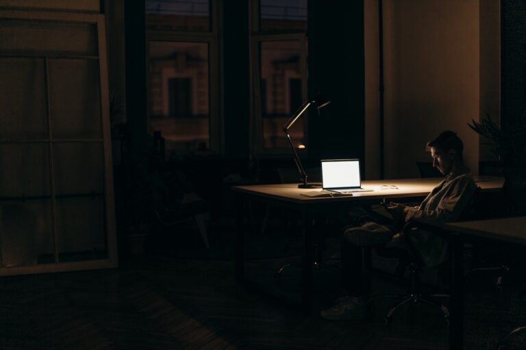 A person sitting at a desk in the dark and looking at their phone. There is a laptop screen open in front of them.