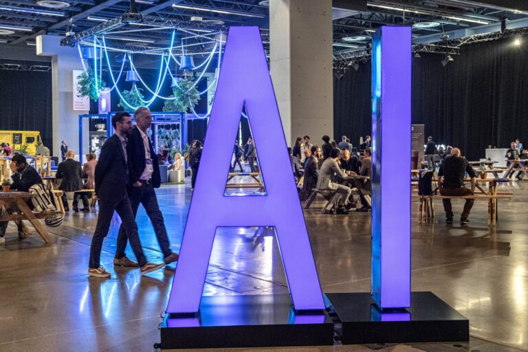 Large purple letters spell out “AI” on a display at the All In AI conference in Montreal. Two men wearing blazers walk by on the left, while other attendees sit at tables and tree-like decorations hang from the ceiling in the background.