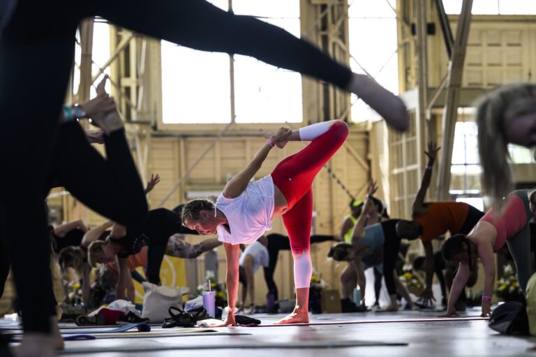 A person in red leggings holds their ankle behind their back with their palm flat on the ground in a yoga pose.