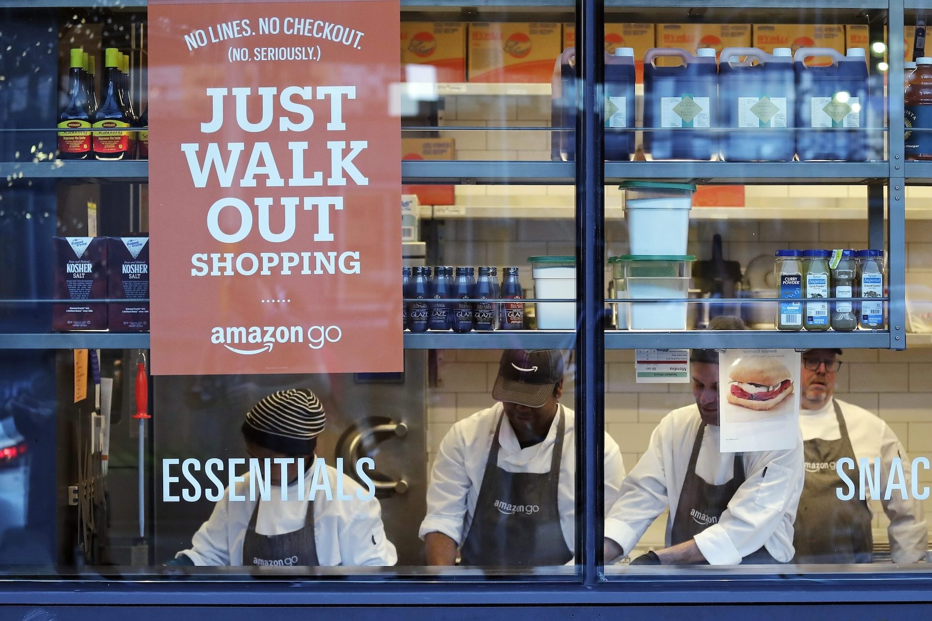 4 workers are seen in a food store from a window in white shirts and black Amazon Go aprons. A sign on top left of the window says “No lines. No checkout. (No. Seriously.) Just Walk Out shopping.