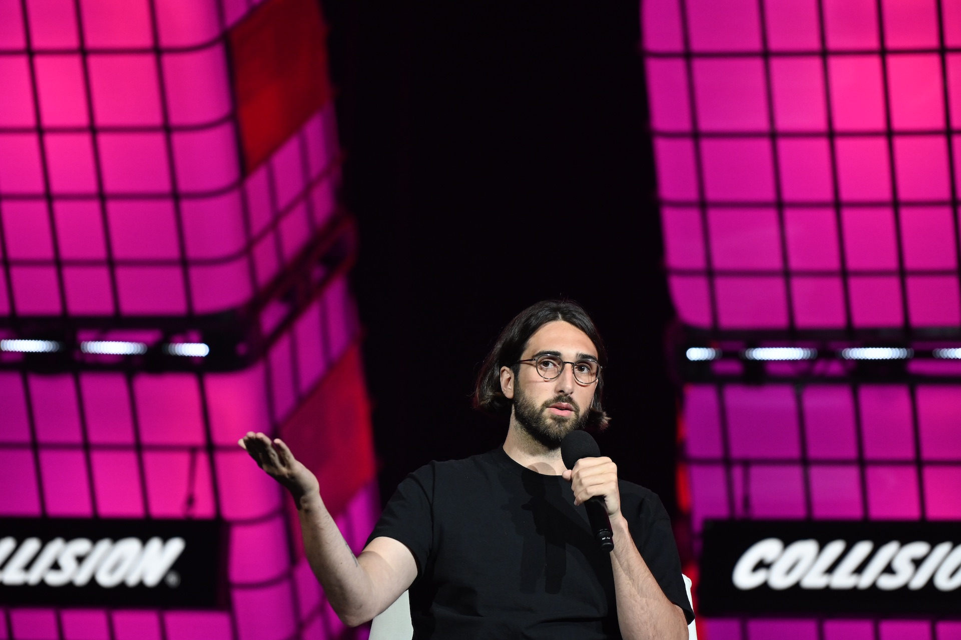 Cohere CEO Aidan Gomez holding a microphone in one hand and raising the other hand forward. The backdrop is pink and black with signs for the Collision technology conference on it.