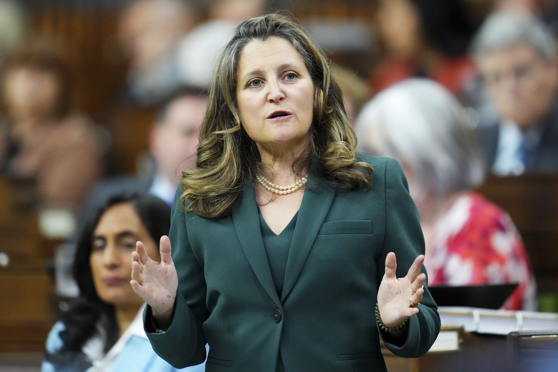 Chrystia Freeland stands with her hands gesturing open. She wears a green suit and a pearl necklace.