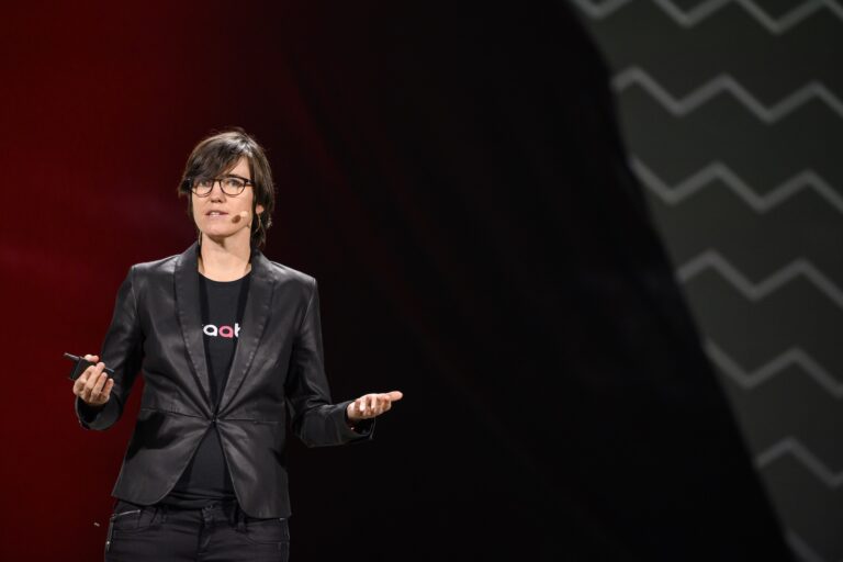 Waabi founder and CEO Raquel Urtasun in a black leather jacket and spectacles. She is standing and speaking into a microphone that is attached to her ear.