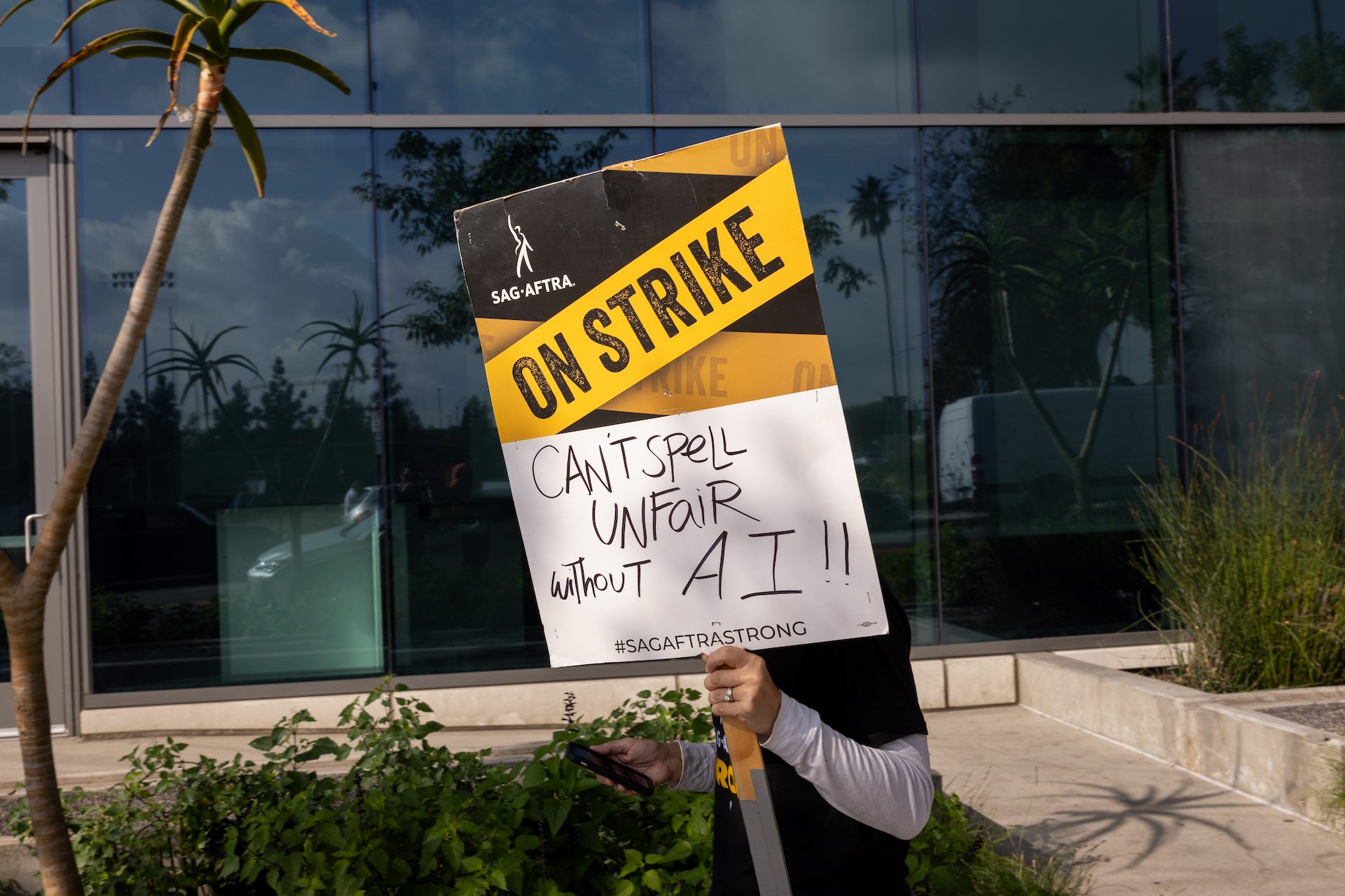 A person holds a SAG-AFTRA sign that says, “Can’t spell unfair without AI.” Their face is blocked by the sign.