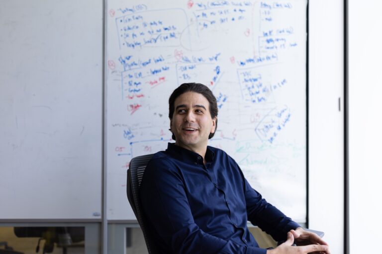 Photo of WonderFi CEO Dean Skurka in a dark blue shirt, sitting in a chair. There’s a whiteboard in the background with some writing on it.