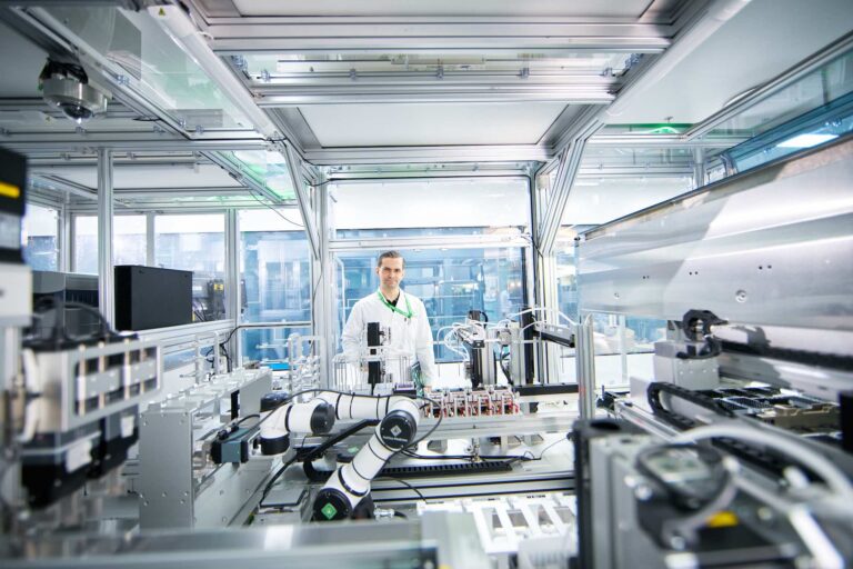 Insilico Medicine founder and CEO Alex Zhavoronkov is seen standing. He is wearing a labcoat and spectacles, and is surrounded by AI-powered robotic lab equipment.