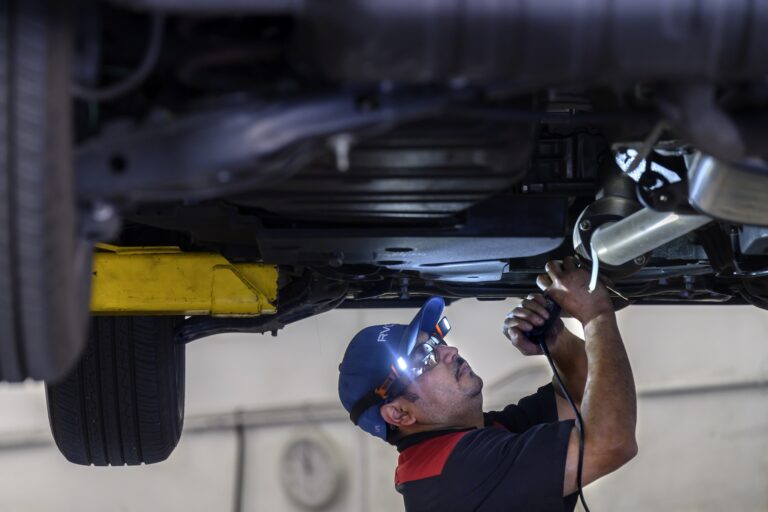 An automotive technician is seen under a car, attaching a catalytic converter to the bottom of it with both hands.