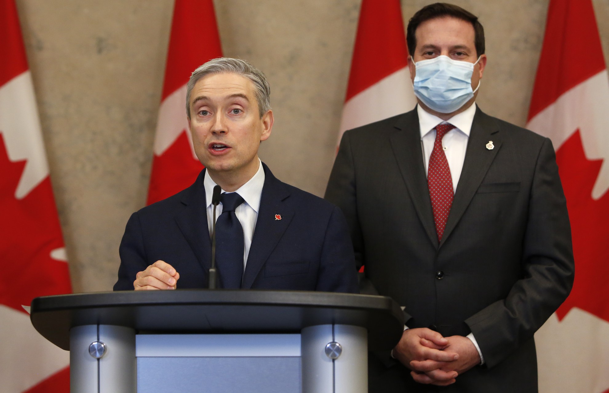 Innovation Minister François-Philippe stands at the podium during a press conference. Public Safety Minister Marco Mendicino is in the background, wearing a mask.