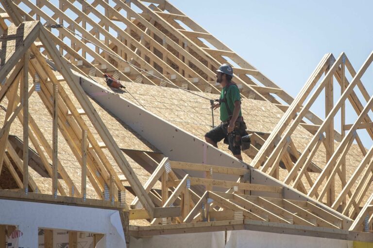 A construction stands on the roof of a house being built out of wood. He wears a green t-shirt and a grey hard hat.