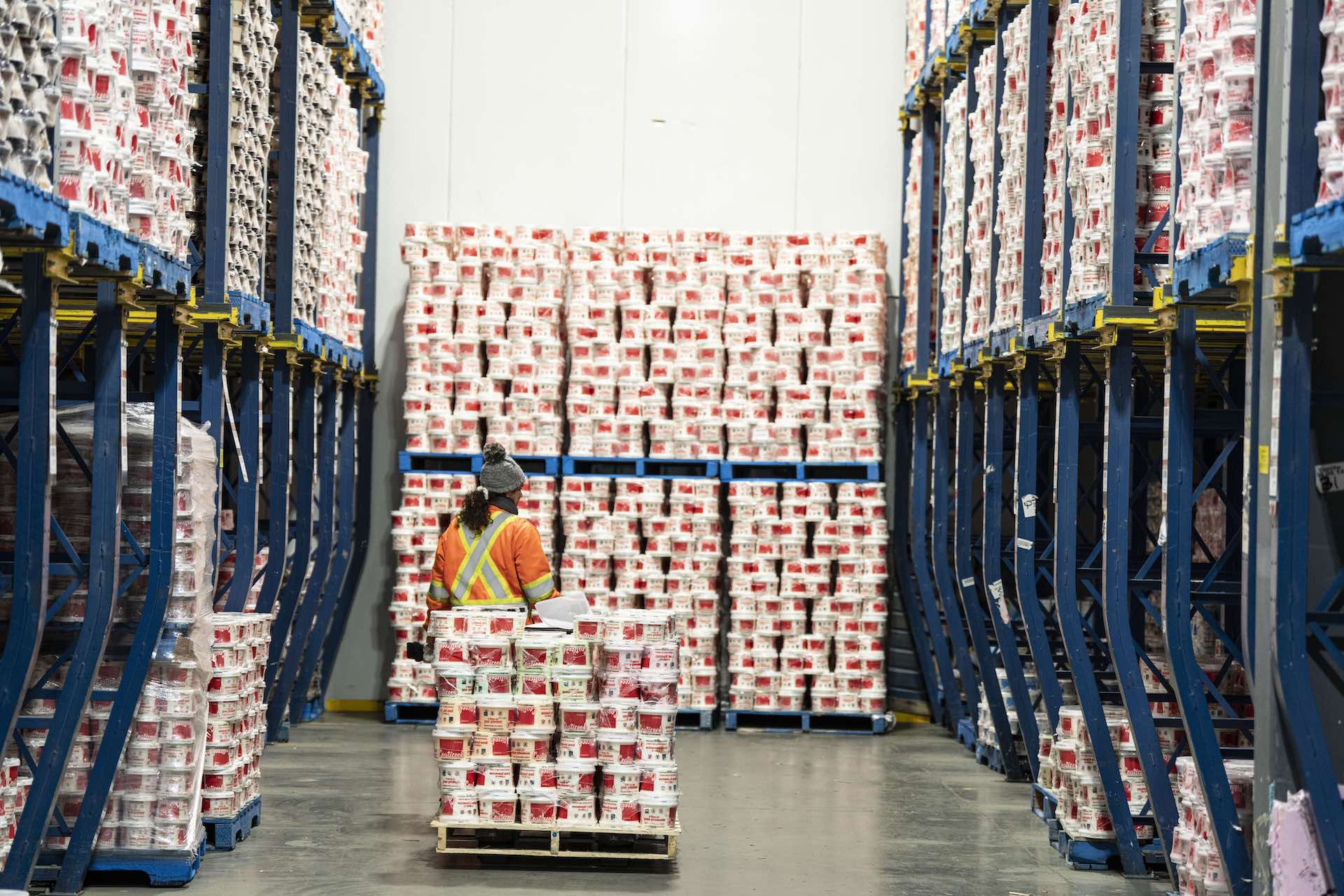 A worker in an orange vest and a grey hat with a pallet of red and white ice cream tubs, surrounded by wall-to-wall pallets of tubs.