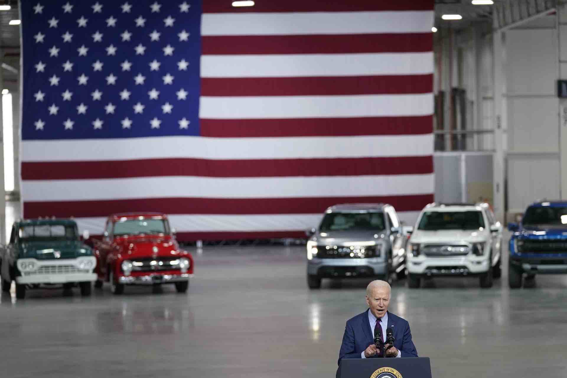 President Joe Biden speaks in front of Ford vehicles. A large U.S. flag is behind him.