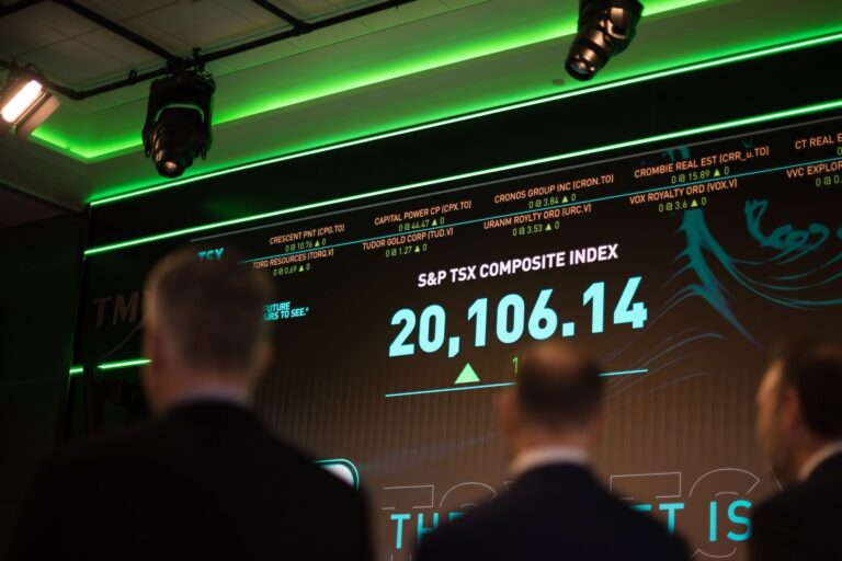 Three people wearing suits look at the S&P TSX Composite Index on the screen.