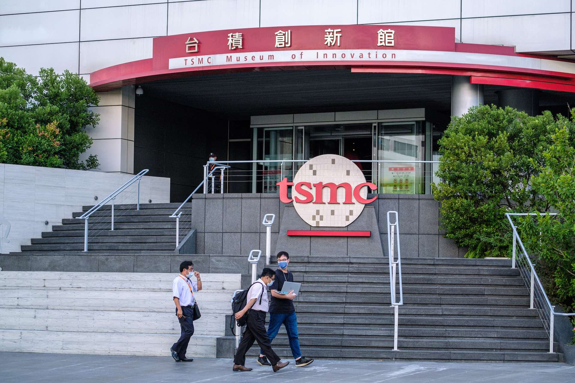 Three men walk by a building with the Taiwan Semiconductor Manufacturing Company logo on it. The logo is a gold sphere with the letters TSMC across the front in lowercase.