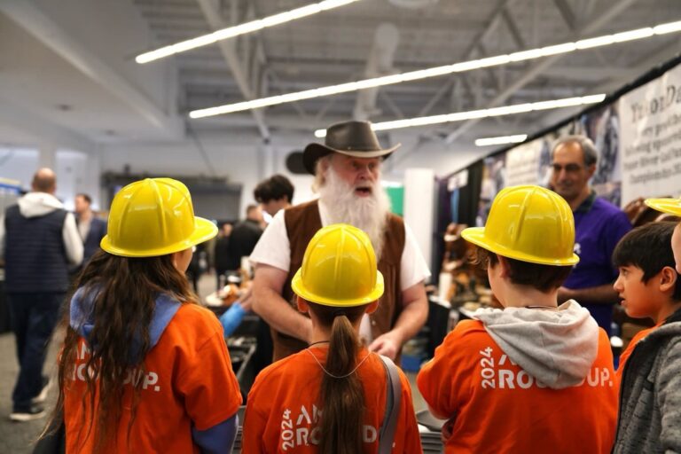 Children wearing orange vests and yellow hard hats listen to a person with a beard and brown hat in the exhibit hall of the Vancouver Convention Centre in January 2024.
