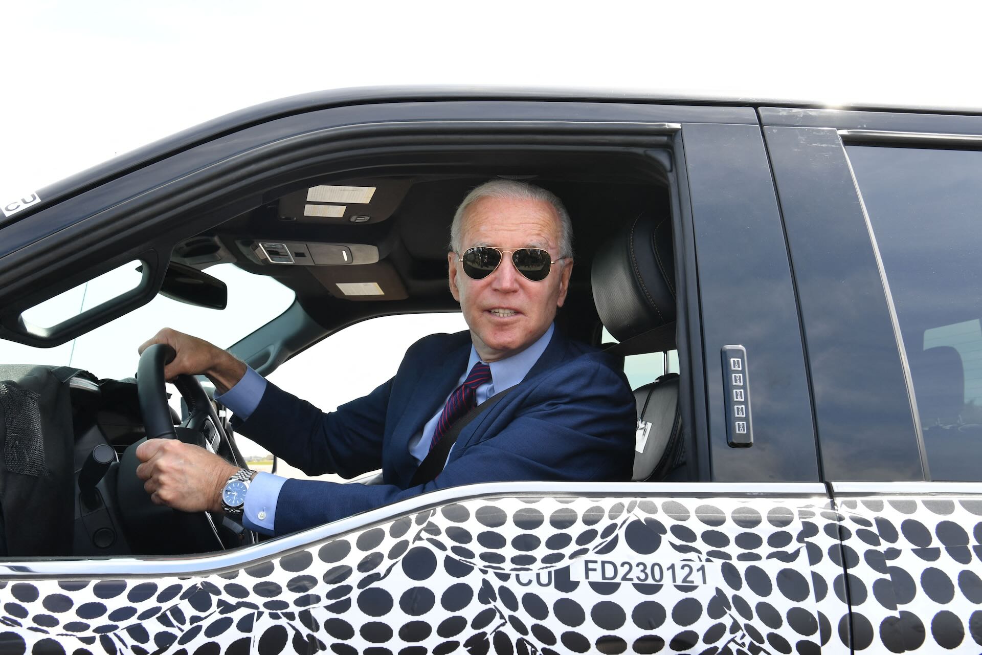 U.S. President Joe Biden looks out the window from the drivers seat of an electric Ford F-150. He wears aviator sunglasses.