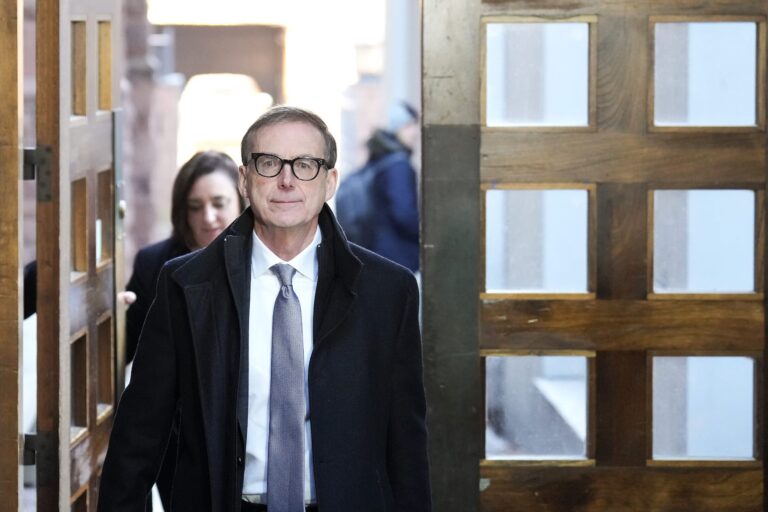 Tiff Macklem walks through a set of wooden doors. He wears a suit and black-framed glasses.