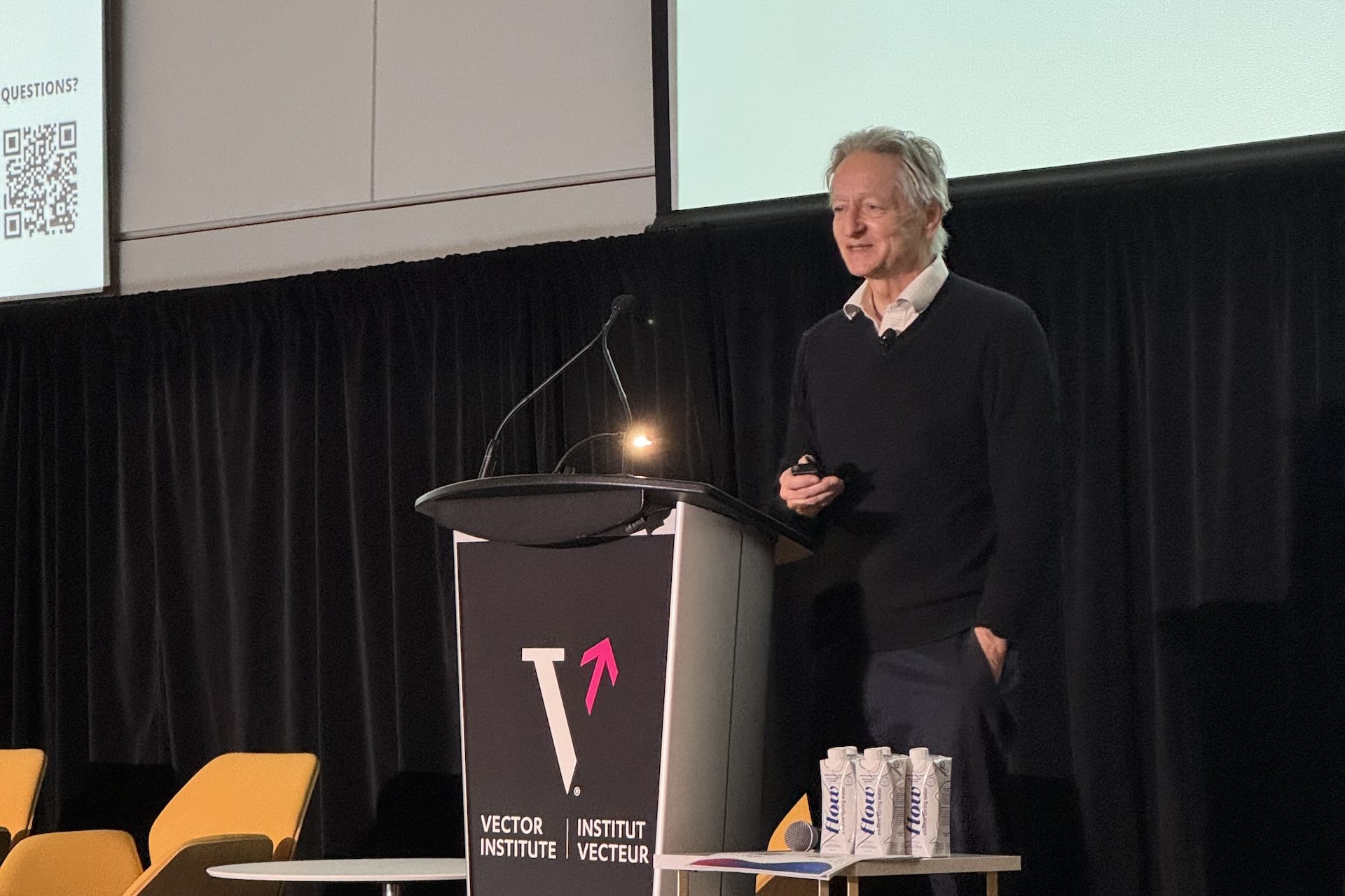 AI godfather Geoffrey Hinton stands at a podium that says “Vector Institute.” He is holding a clicker in one hand and has his other hand in his pocket.