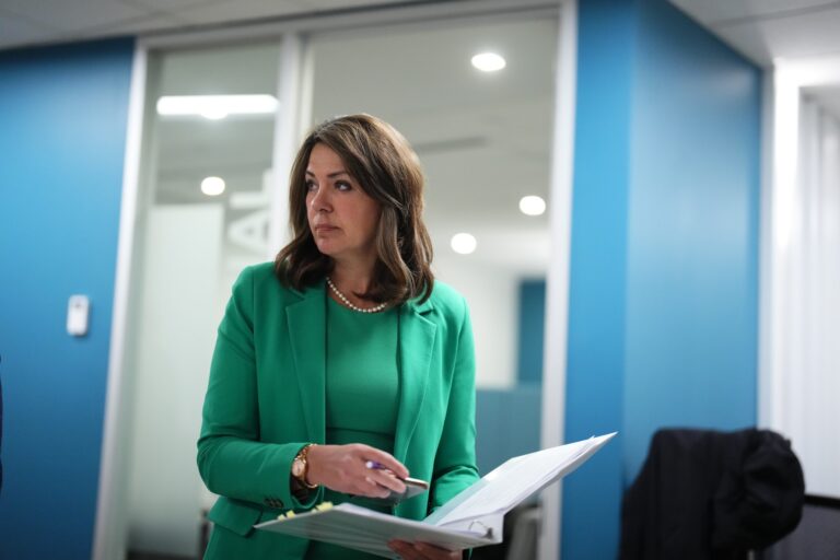 Alberta Premier Danielle Smith wears a green suit and a string of pearls. She holds an open binder in her left hand.