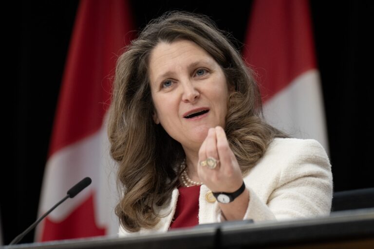 Finance Minister Chrystia Freeland wearing a red top and white blazer. She is gesturing with her left hand and there are blurred-out Canadian flags in the background.
