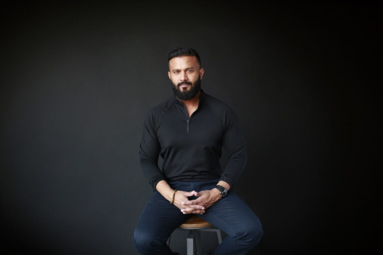 Lightspeed founder and interim CEO Dax Dasilva sitting on a stool in a black shirt and blue pants. He has his fingers interlocked, resting on his lap.