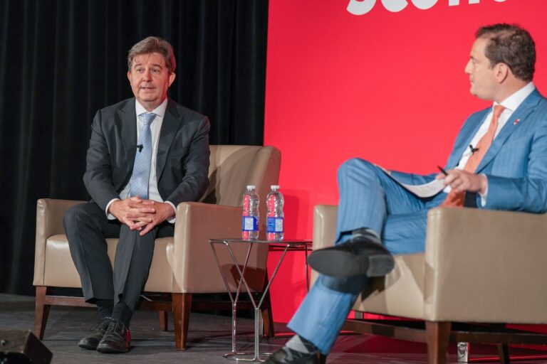OSFI superintendent Peter Routledge, wearing a black suit, sits on a chair and speaks to a panel moderator wearing a blue suit.