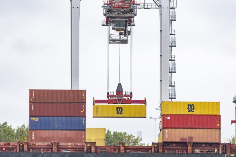 Alt: A yellow shipping container is being loaded onto a container ship with red, blue, orange, and yellow-coloured containers.