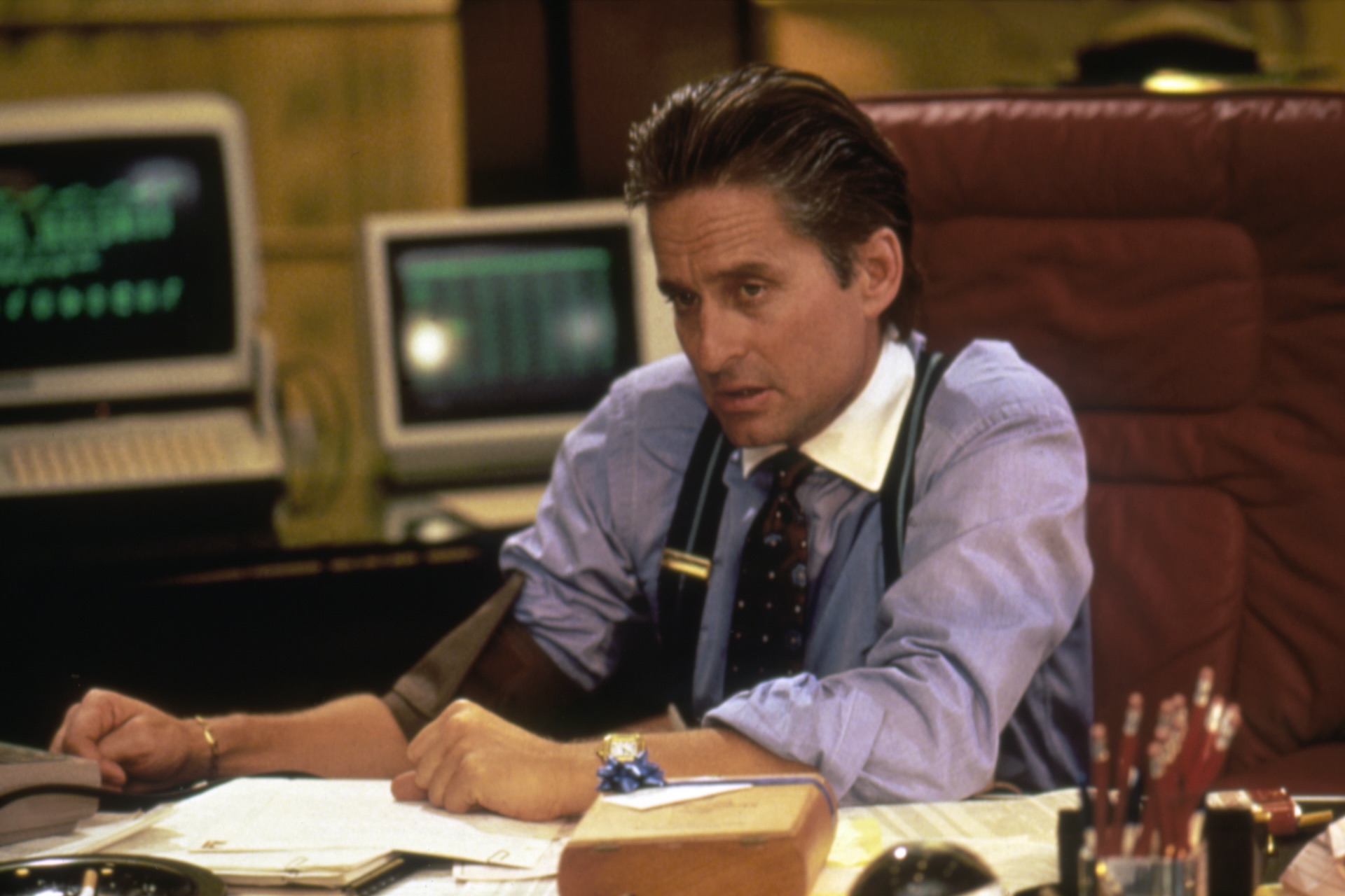 Gordon Gekko, a character played by Michael Douglas in the film Wall Street, sits at a desk with papers strewn about. He has greased-back brown hair and wears a blue button-down shirt, a reddish-brown tie, suspenders and a gold watch. Computers showing stock prices are in the background.