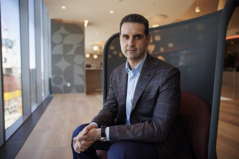 Abraham Tachjian sits in large-backed black chair, angled slightly toward a window. His hands are clasped, and he wears a blue shirt with a brown blazer.