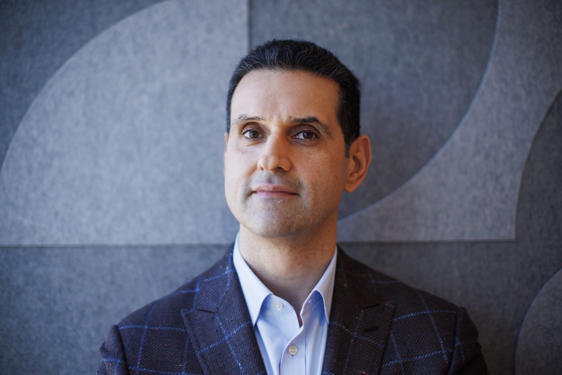 Abraham Tachjian is pictured from the chest up, slightly smiling. He wears a blue shirt and brown blazer with blue square outlines on it.