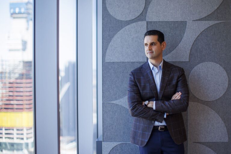 Abraham Tachjian stands with his arms crossed to the right of a window. He wears a blue shirt and a brown blazer with blue square outlines on it.