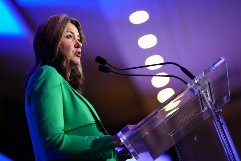 Alberta Premier Danielle Smith speaking into a microphone at a translucent podium. She is wearing a green blazer.