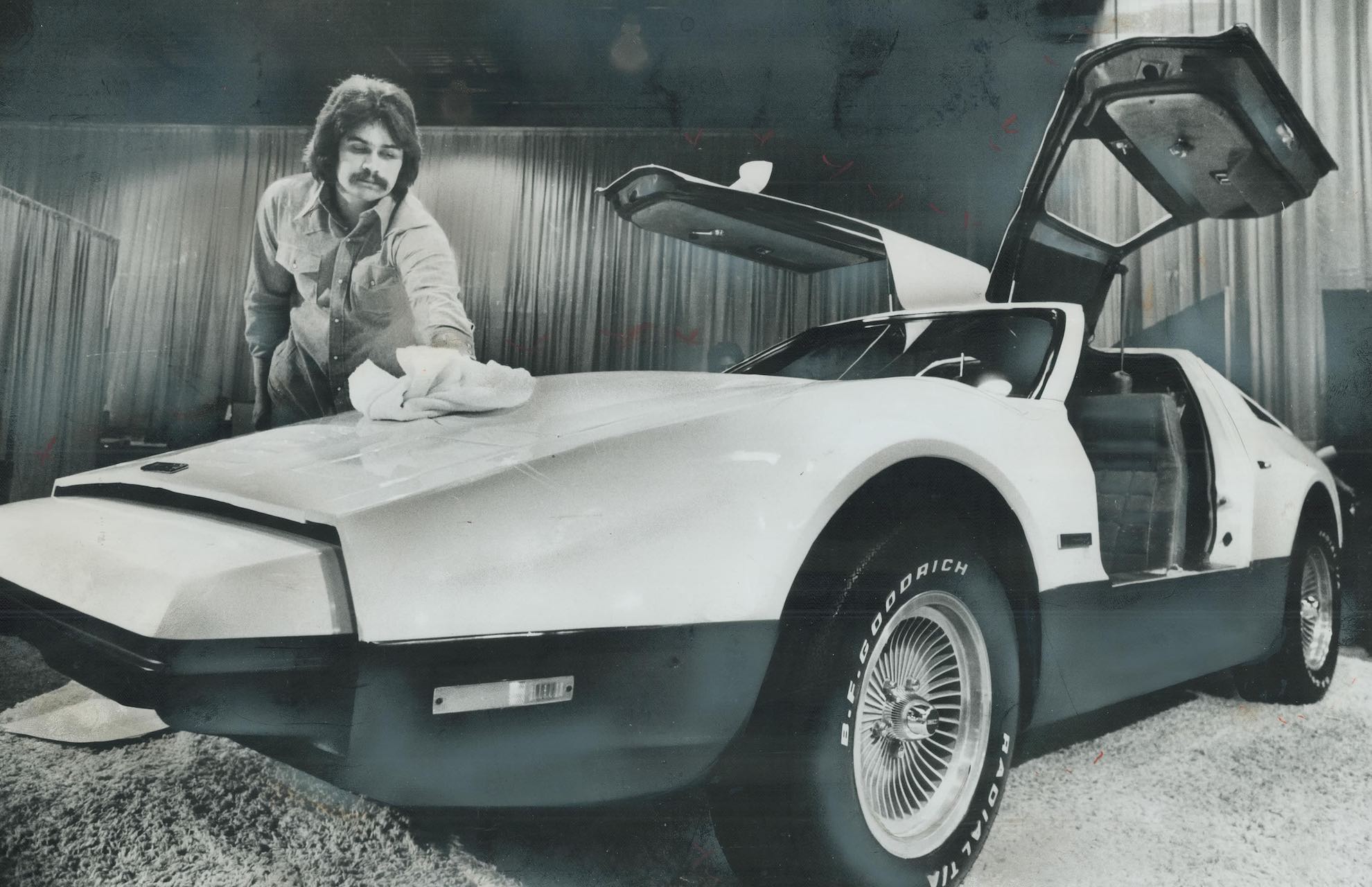A man polishes a 1975 Bricklin model with the doors open. He has long hair and a moustache.