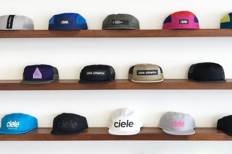 A colourful selection of Ciele running hats are displayed on three wooden shelves against the backdrop of a white wall.