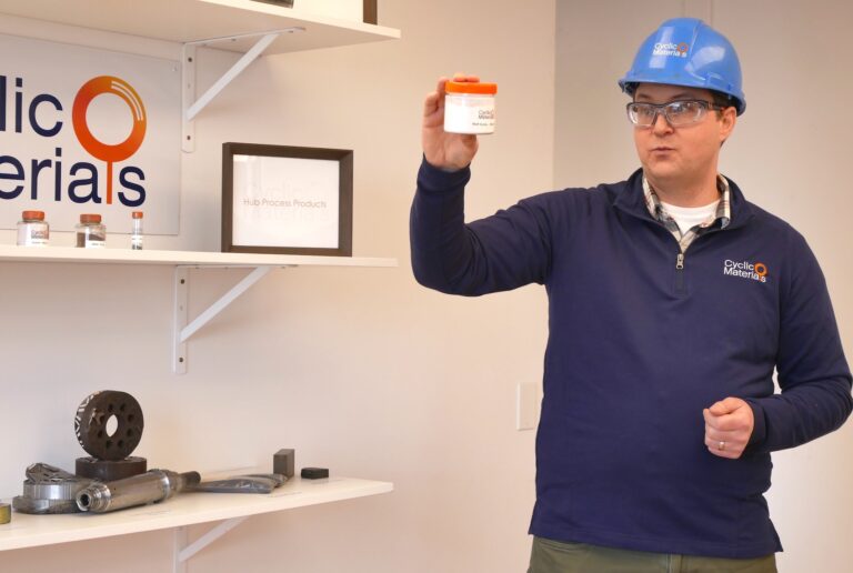 A man in a blue hardhat and blue quarter zip holds a jar of white powder. He wears safety glasses and stands to the right of a Cyclic Materials sign.