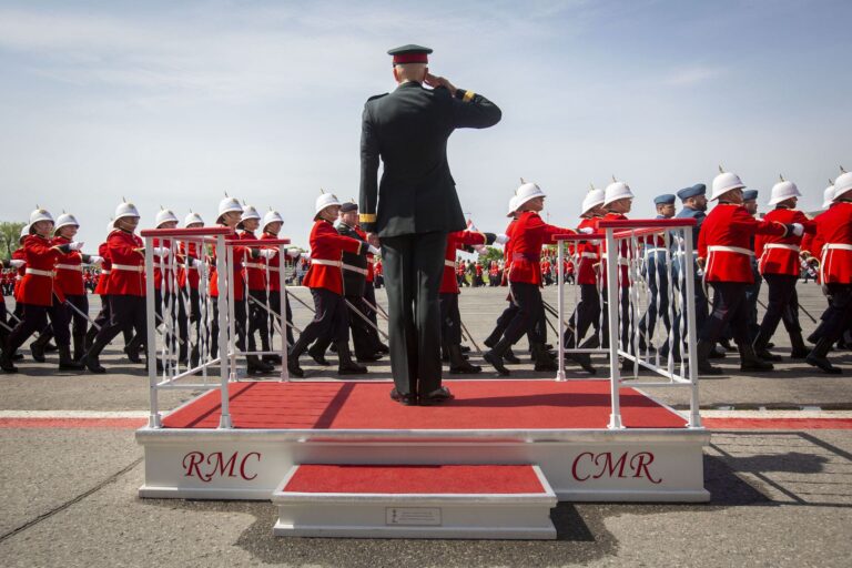 A military commander in full-dress uniform stands on a dais with his back to the camera and salutes as a class of newly graduated officers wearing red coats marches past.