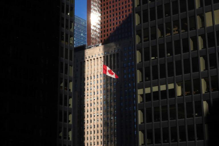 A red-and-white Canadian flag flies in front of tall glass office buildings in Toronto’s financial district.