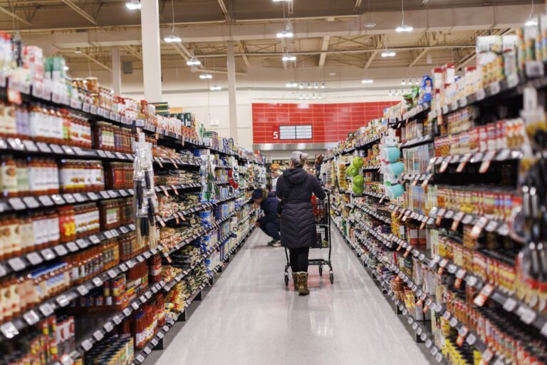 A woman stands with her back facing the camera pushing a grocery cart in an aisle of pasta sauce and canned goods. She wears a black coat and brown boots.