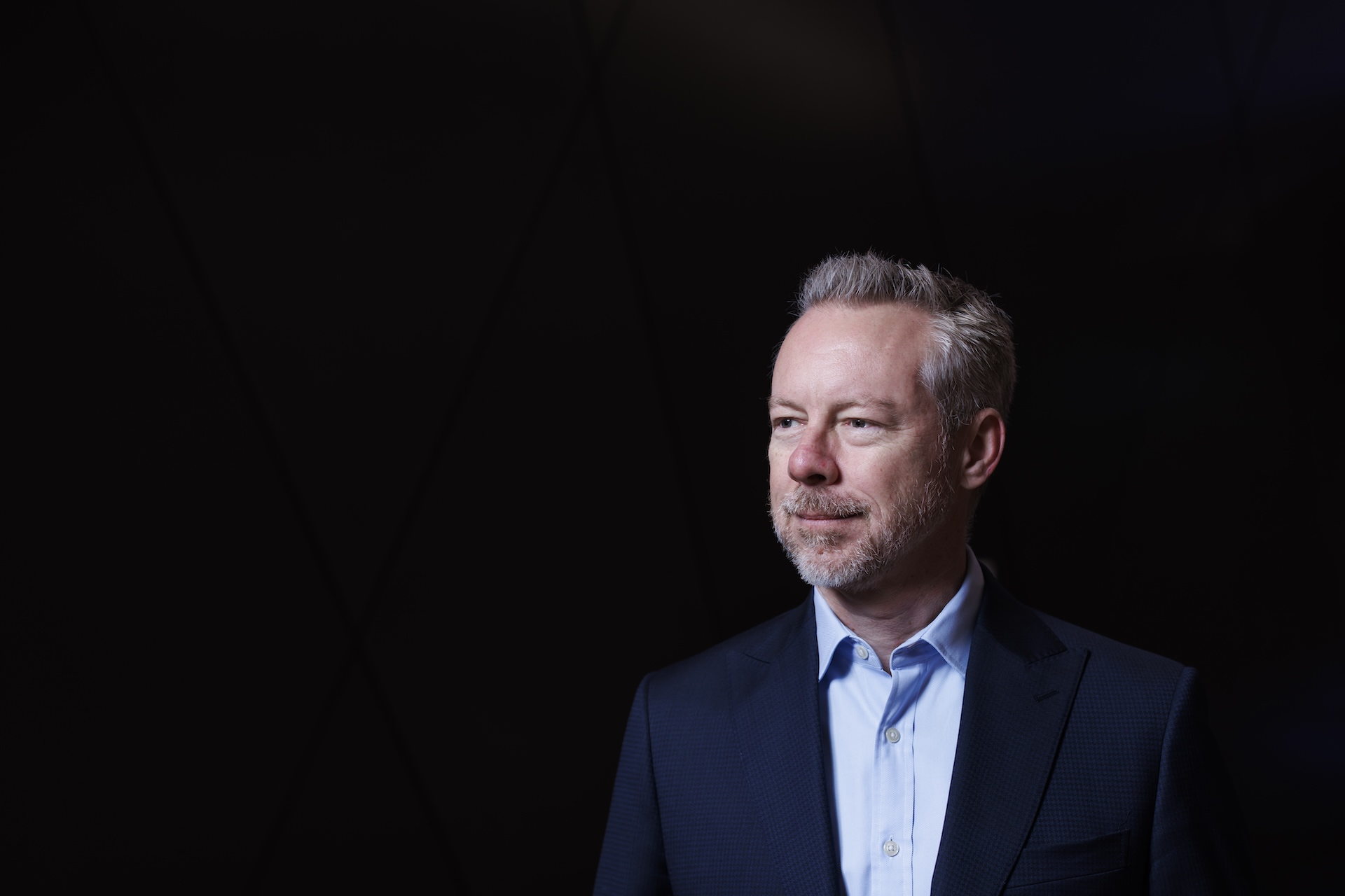 John McKenzie is pictured gazing to the left, in the third right section of this image. He has grey hair, and wears a light blue shirt with a dark blue blazer.