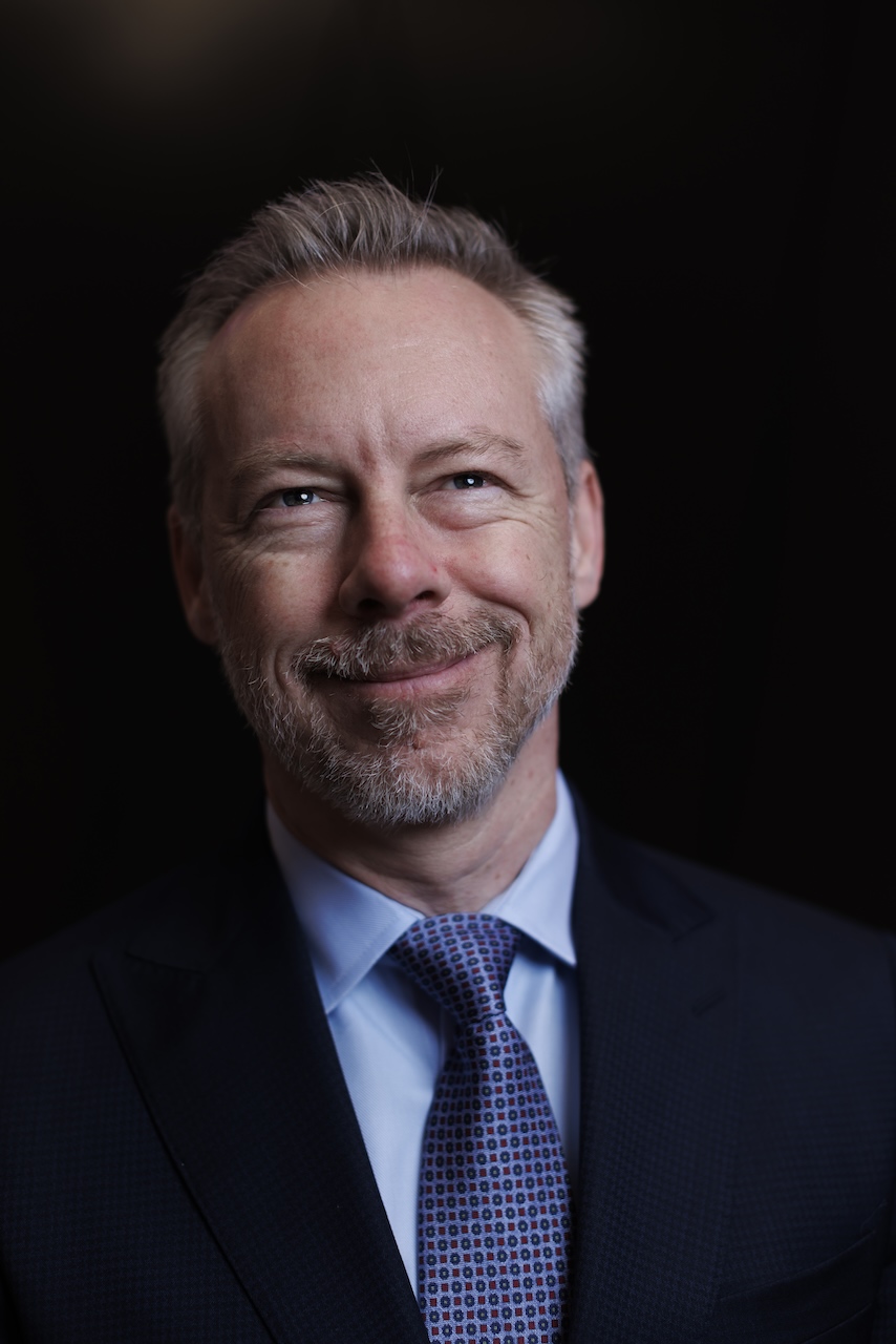 A close-up portrait of TMX Group CEO John McKenzie wearing a dark blue suit and blue-and-red spotted tie.
