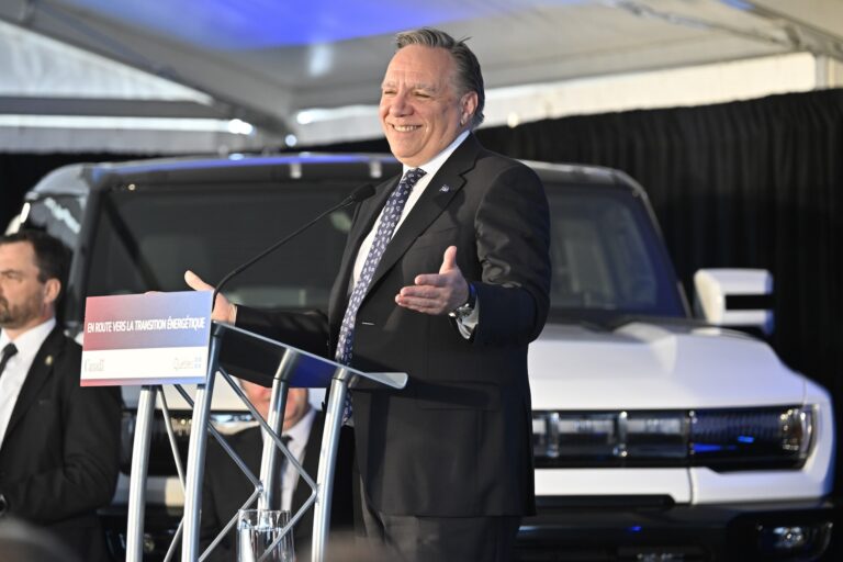 Quebec Premier François Legault appears at a lectern with a white EV in the background. Legault, who is wearing a dark suit, is smiling and has his arms slightly raised in a shrugging motion.
